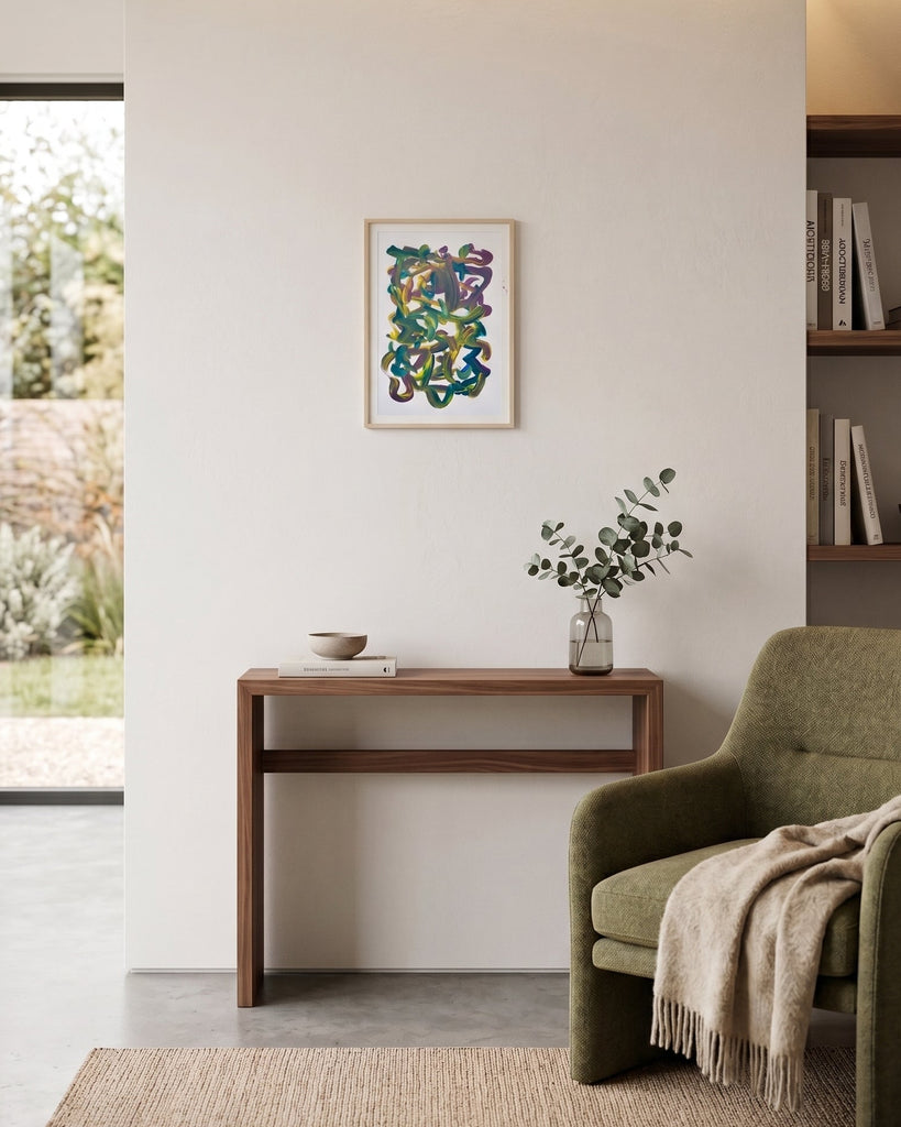 A framed abstract art with colorful squiggly lines hangs on a white wall in a modern, sunlit living space. Below the artwork is a wooden console table with a small vase of greenery, and to the right is an olive green armchair with a beige throw blanket.