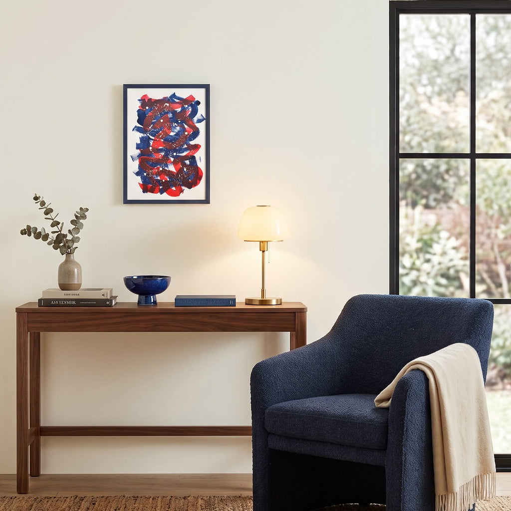 A styled interior scene with a navy upholstered armchair beside a wooden console table, a lit table lamp, books, a vase with dried stems, and a framed abstract artwork on the wall. A window with black mullions is visible on the right.