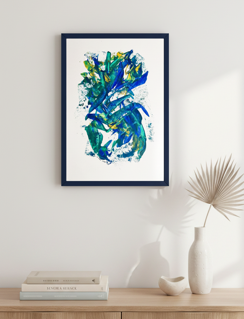 Framed abstract wall art hanging on a light wall above a wooden console, with stacked books, a small bowl, and a dried palm leaf in a vase. The artwork features blue, teal, green, and yellow brushstrokes on a white background with a dark navy frame.