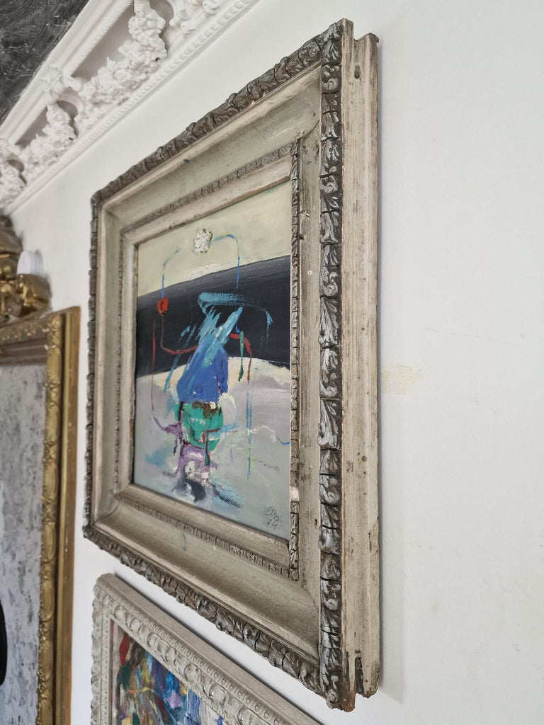 An abstract landscape painting in a distressed, ornate silver frame, hanging on a white wall next to other framed art, side view.