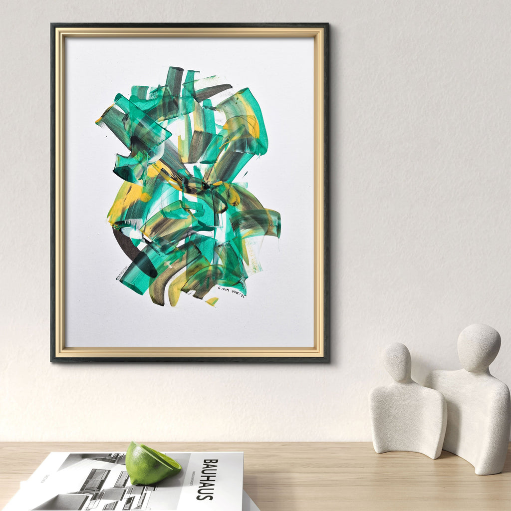 Framed abstract art piece on a wall above a wooden surface with magazines and sculptures.