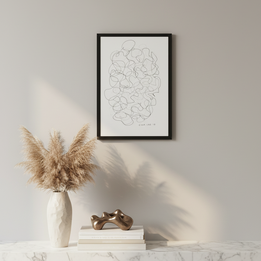 A minimalist framed art with a black line drawing, styled on a white marble shelf with a vase of pampas grass and a small bronze sculpture against a warm gray wall.