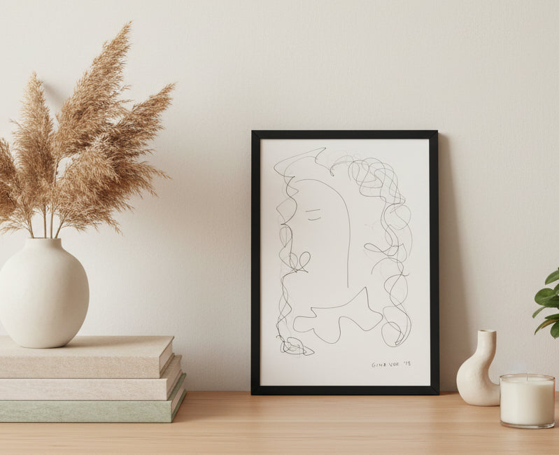 A framed minimalist line art featuring a ghostly face with curly hair, signed 'Gina Voss 19' in the lower right corner, displayed against a light gray wall.