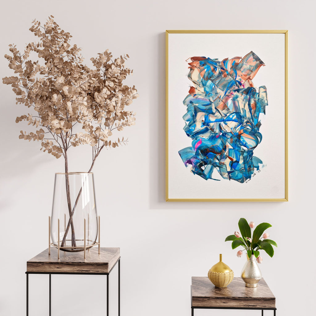 Abstract art piece in a gold frame on a wall with decorative plants and tables.