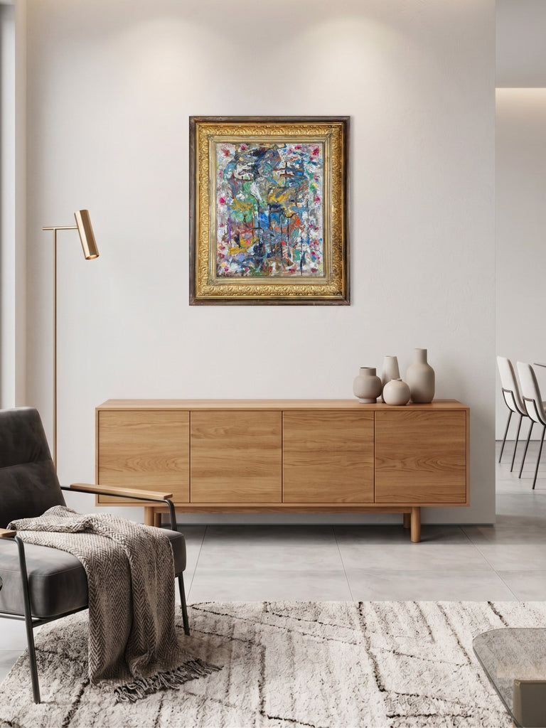 A colorful abstract painting in a gold frame hangs on a light gray wall above a mid-century modern wooden credenza in a styled living room setting.