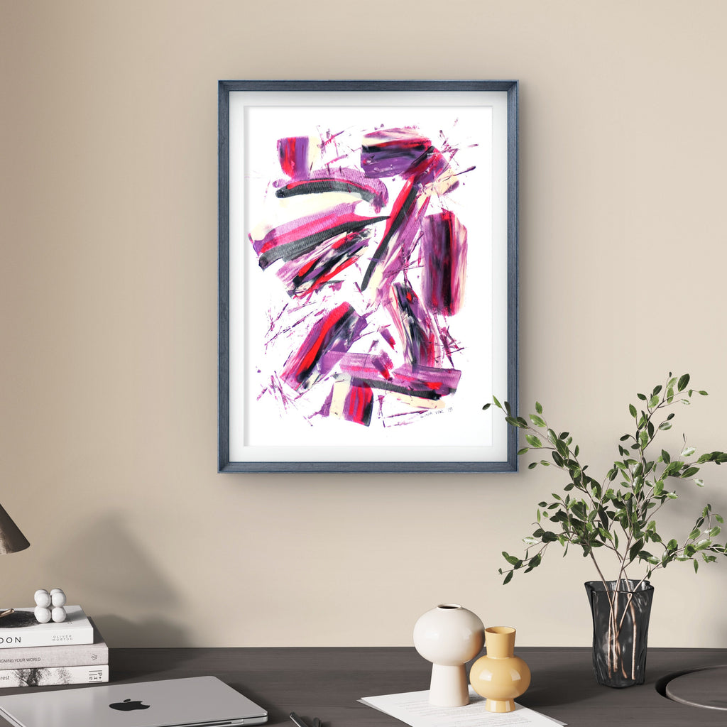 Framed abstract art piece on a wall above a desk with a laptop and decor items.