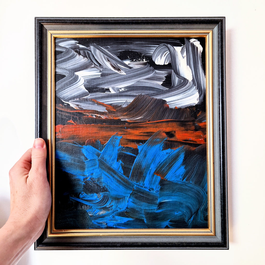 Abstract painting with blue and orange strokes in a black frame held by a hand.